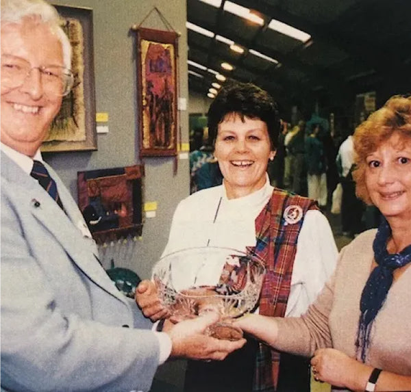 Iain MacPherson and two ladies shaking hands and smiling at a textile exhibition.